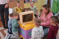 7. Na ulu edukacyjnym stoi ramka z planszą dydaktyczną przedstawiającą społeczność pszczół, wkoło dzieci, nauczycielka.