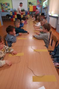 15. Dzieci siedzą przy stolikach, przed nimi plastry wosku, w tle edukator i nauczycielka.