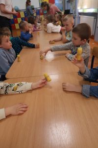 17. Dzieci siedzą przy stolikach, trzymają świeczki z wosku pszczelego wykonane przez nich podczas warsztatów.