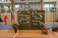 1. Dekoracja uroczystości, kwiaty w wazonie i koszu, trzy flagi białoczerwone, drzewko w koszu wiklinowym.